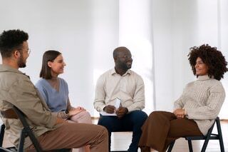 Four people in a circle, seated and talking. A diverse group in a bright room, engaged in a discussion.
