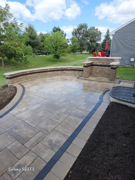 Stone patio with curved retaining wall, built-in fire pit, and garden view beside a house.