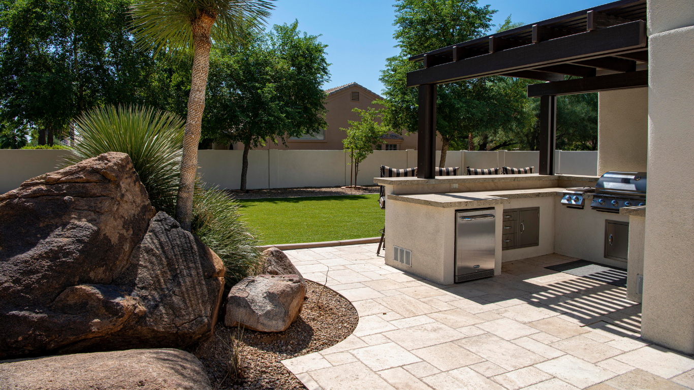 Outdoor patio with stone pavers, built-in grill, and large boulders beside a grassy backyard.