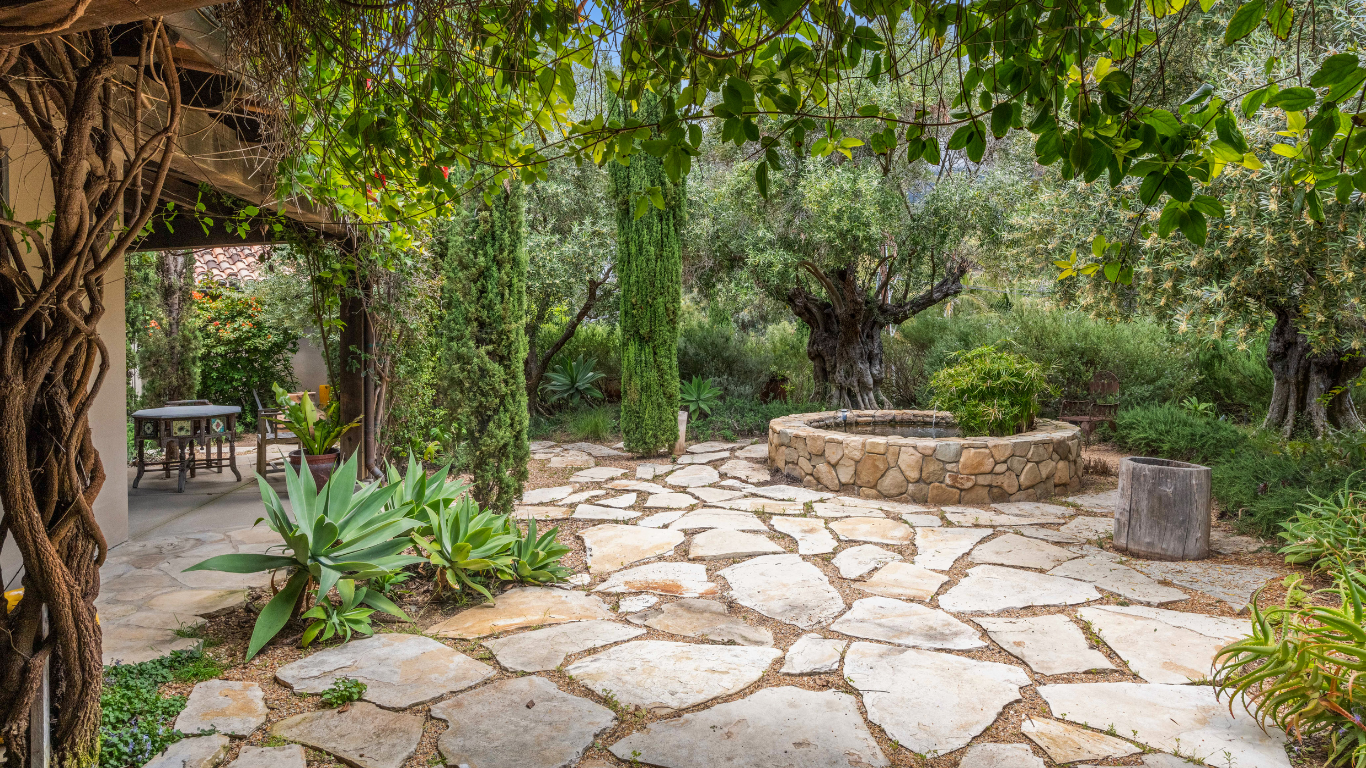 Stone patio path in a lush garden with trees, vines, and outdoor seating under a shaded pergola.