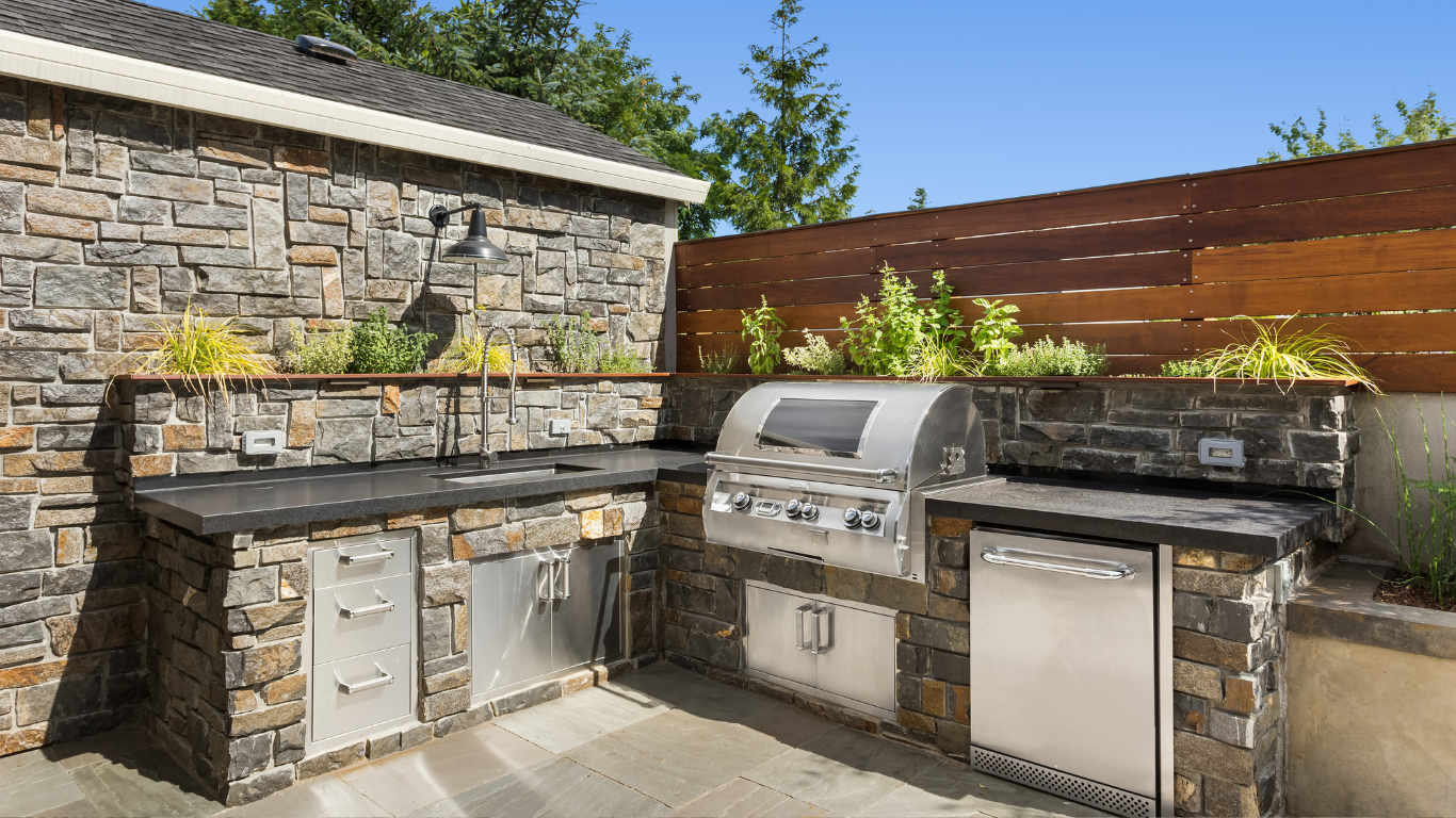 Outdoor stone patio with built-in grill, sink, and cabinets against a stone wall and wooden fence