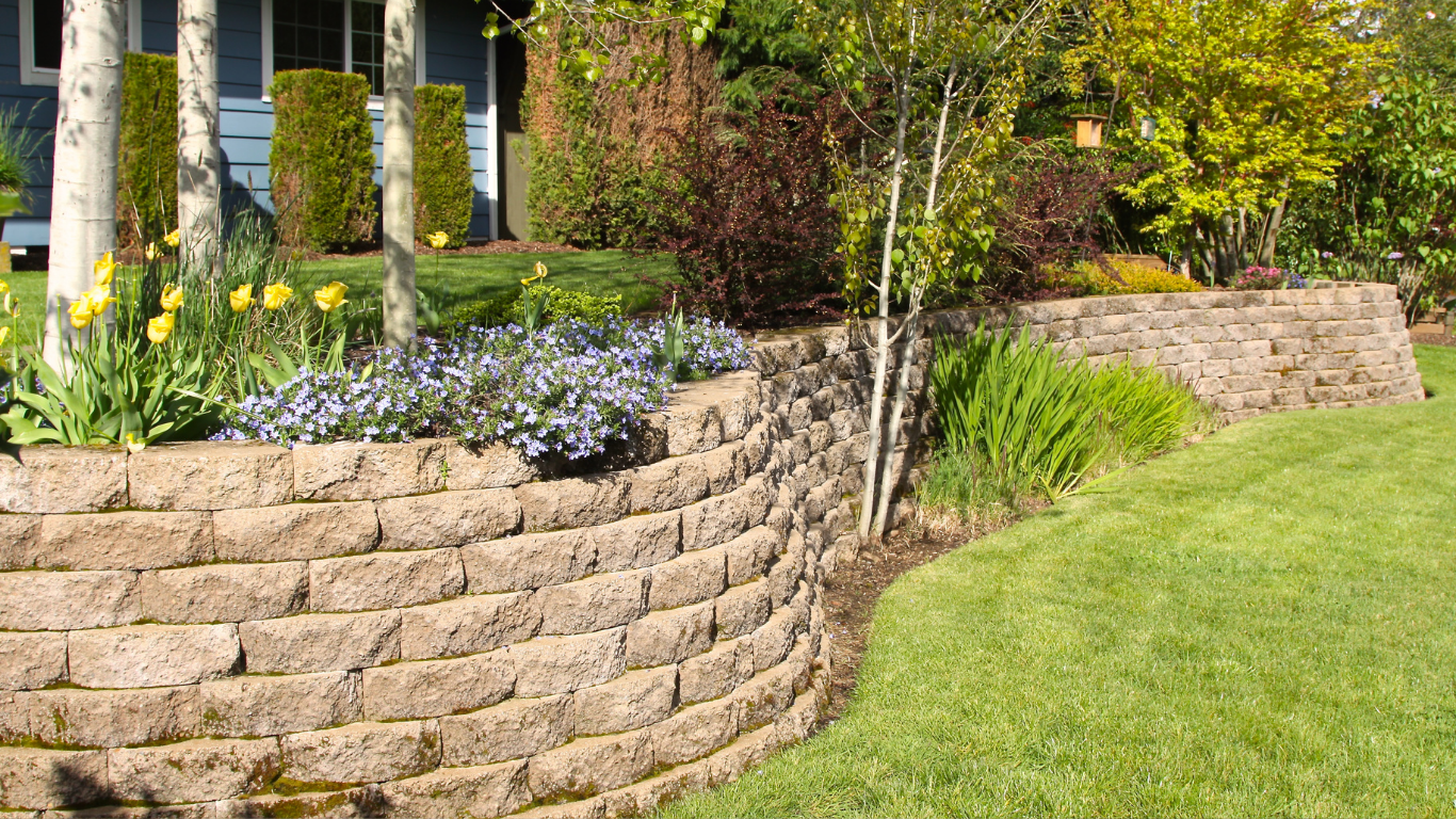 Landscaped garden with stacked stone retaining wall, flowers, shrubs, and green lawn in sunlight