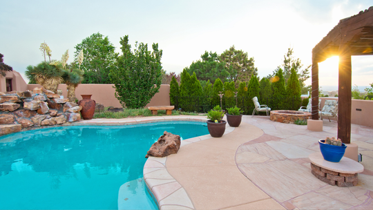 Sunny backyard pool with turquoise water, stone patio, potted plants, and trees at sunset
