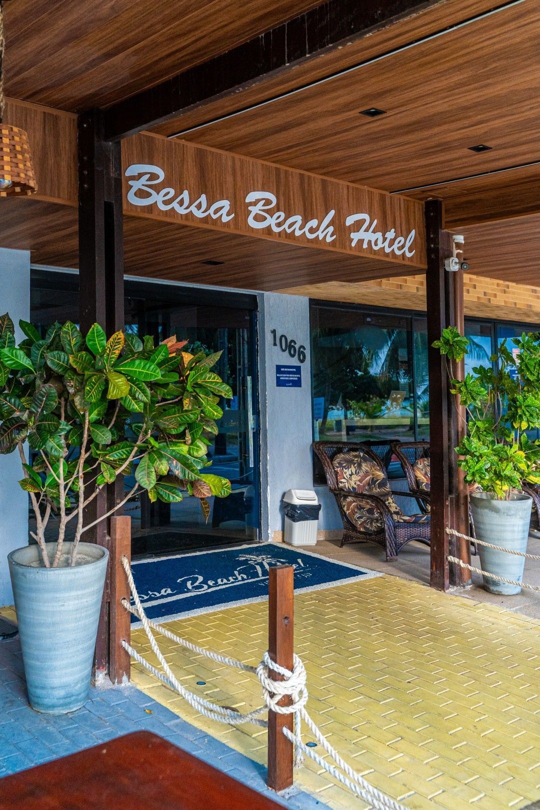 Entrada do Bessa Beach Hotel com cobertura de madeira, vasos de plantas e portas de vidro.