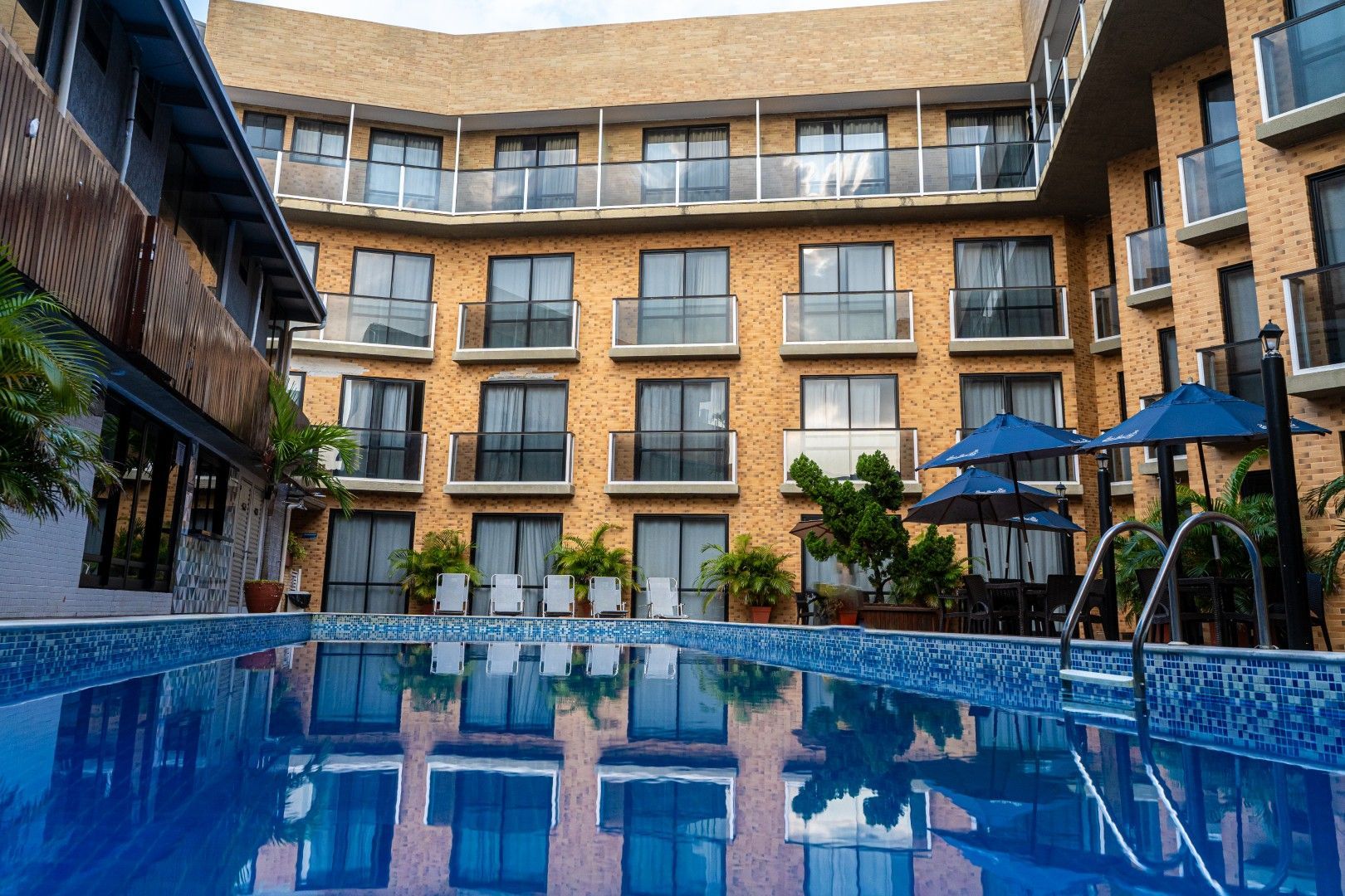 Piscina de hotel com pátio interno, água azul, prédios de tijolos e espreguiçadeiras sob guarda-sóis azuis.