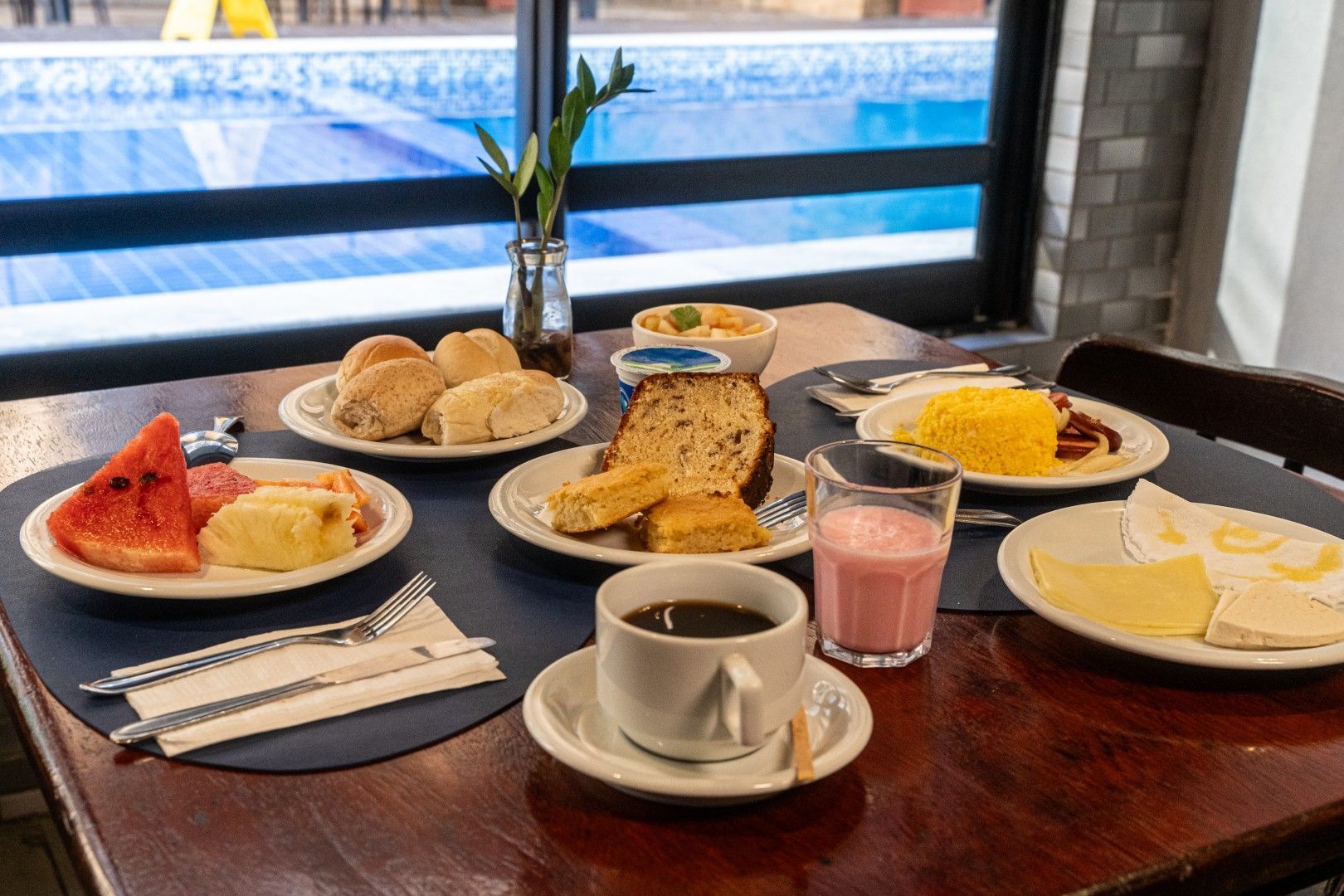 Café da manhã servido em uma mesa de madeira perto da janela, com doces, torradas, ovos, café e um smoothie rosa.