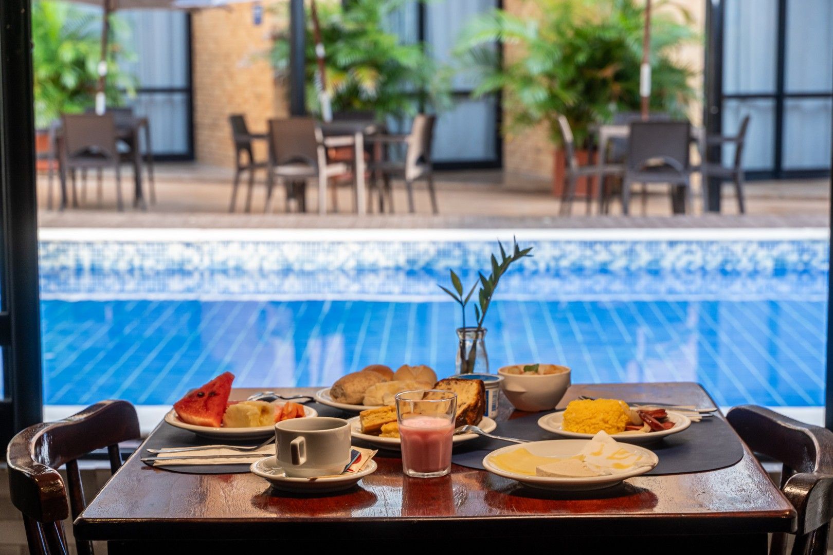 Café da manhã servido em uma mesa à beira da piscina, com torradas, suco e café em um ambiente de resort.