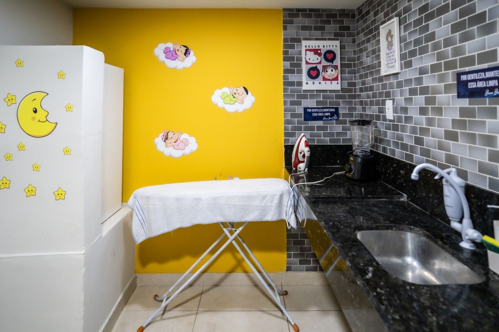 Cozinha pequena com parede de destaque amarela, geladeira branca, tábua de passar roupa e pia com bancada preta.
