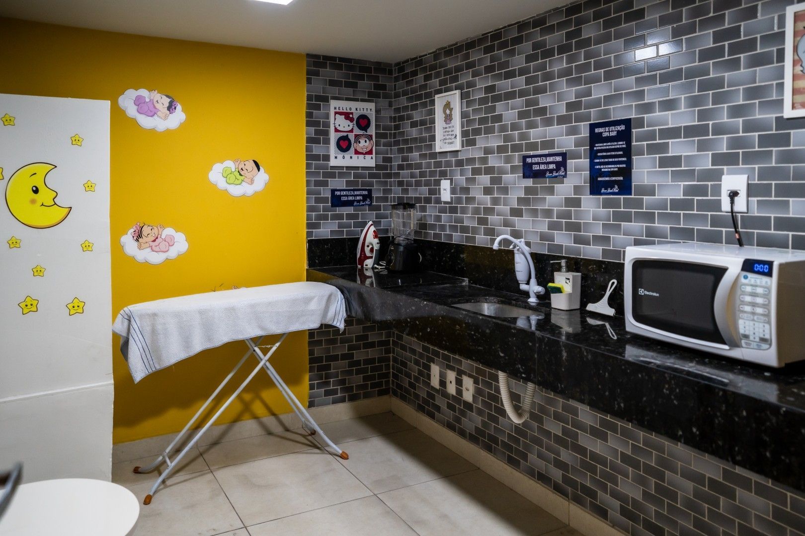 Cozinha compacta e iluminada com parede amarela, tábua de passar roupa branca, pia e micro-ondas sobre uma bancada preta.