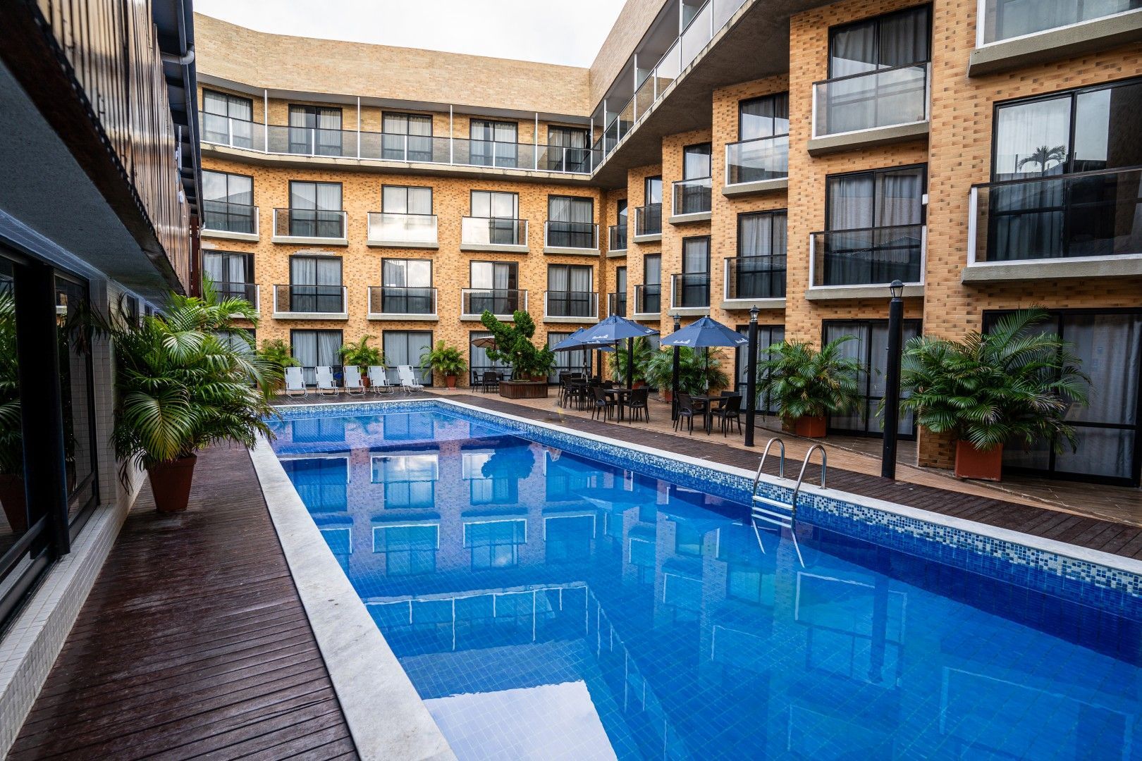 Piscina de hotel com água azul no pátio, rodeada por plantas em vasos e edifícios de tijolos de vários andares.