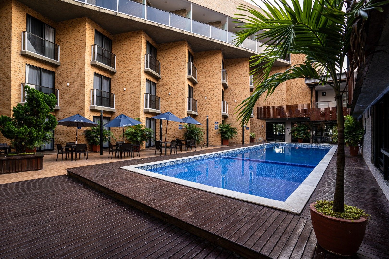 Pátio externo do hotel com piscina azul, edifícios de tijolos e plantas tropicais em vasos.