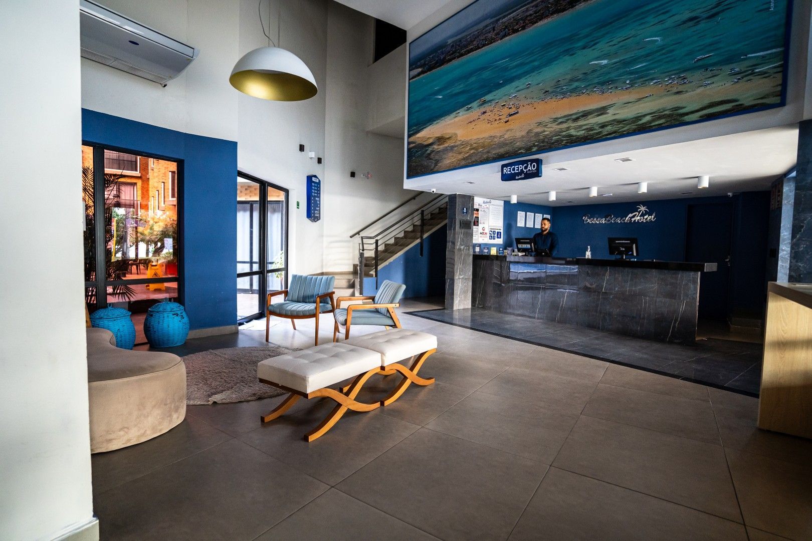 Lobby de hotel moderno com detalhes em azul, balcão de recepção, área de estar e escadaria sob um grande mural.