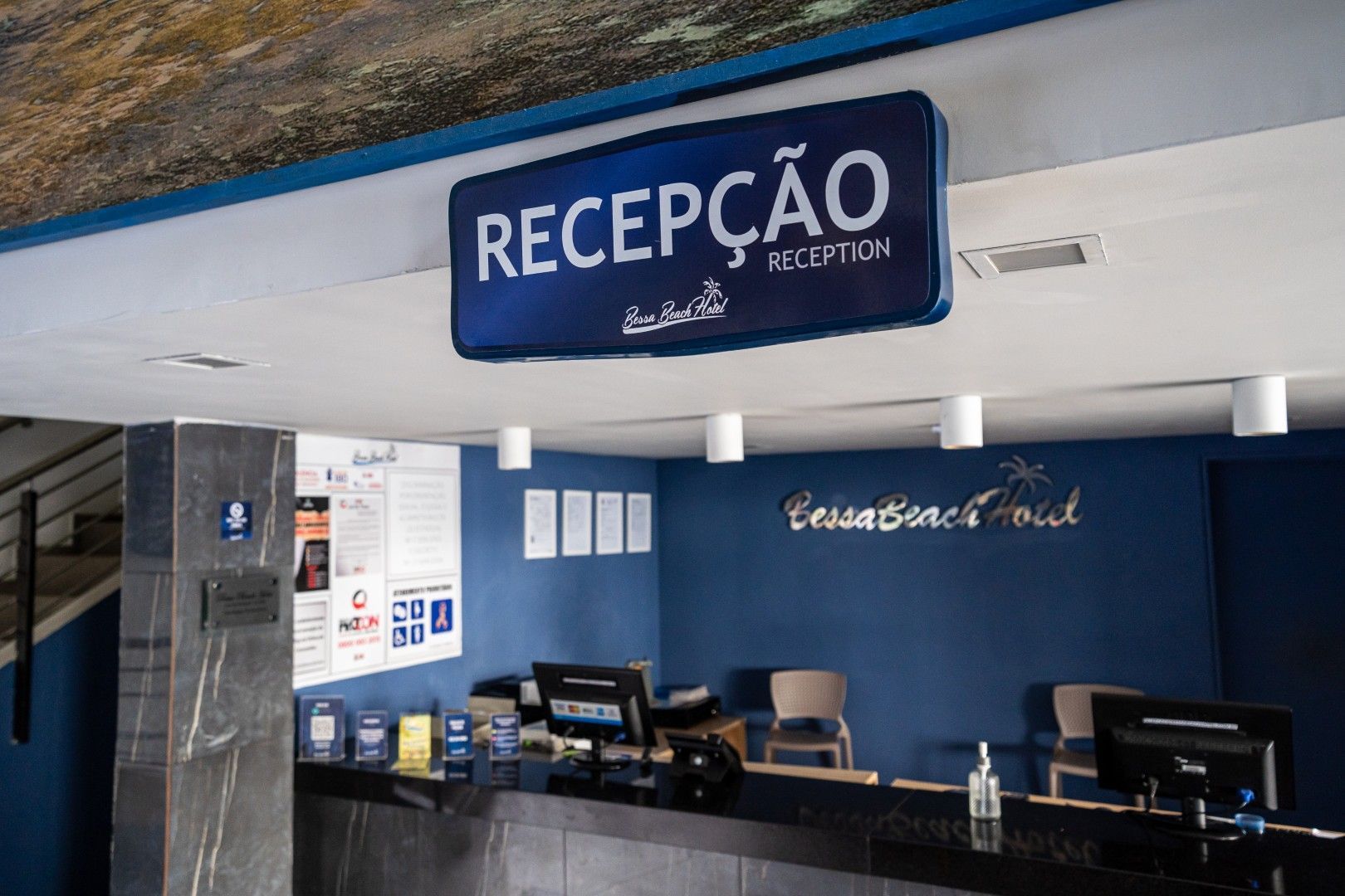 Recepção de hotel com balcão preto, placa azul “RECEPÇÃO” e paredes azul e branca.
