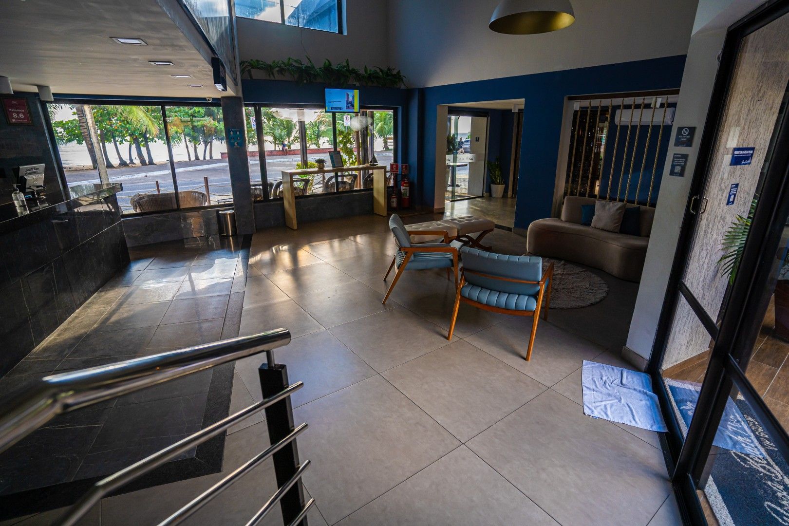 Área de estar moderna no saguão com cadeiras, mesas e piso de azulejos perto da entrada de vidro.