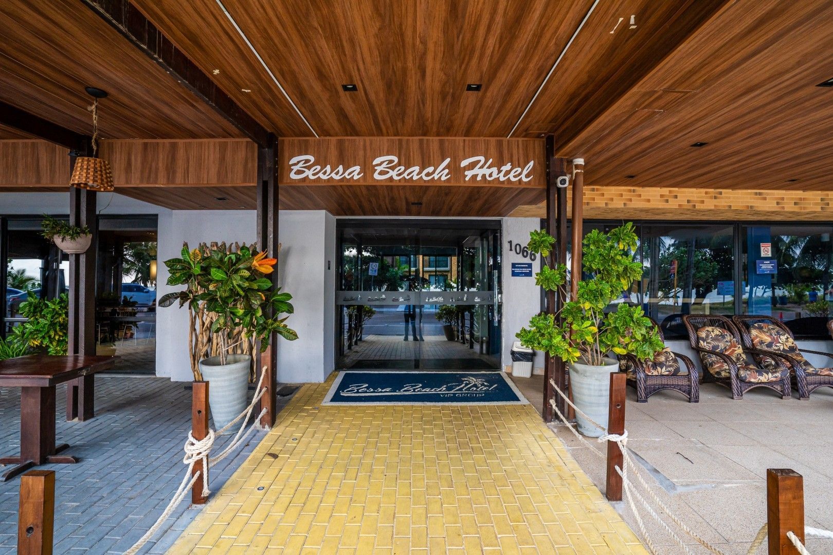 Entrada do Esau Beach Hotel com telhado de madeira, passarela amarela e vasos de plantas.