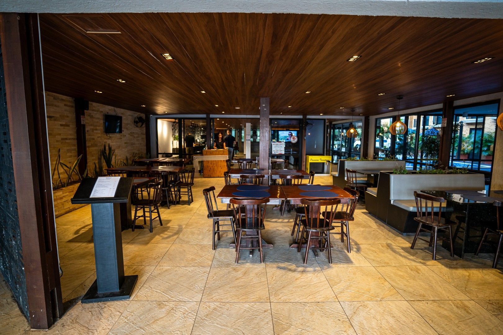 Interior vazio de um restaurante moderno com teto de madeira, piso de azulejo e mesas e cadeiras perto das janelas.