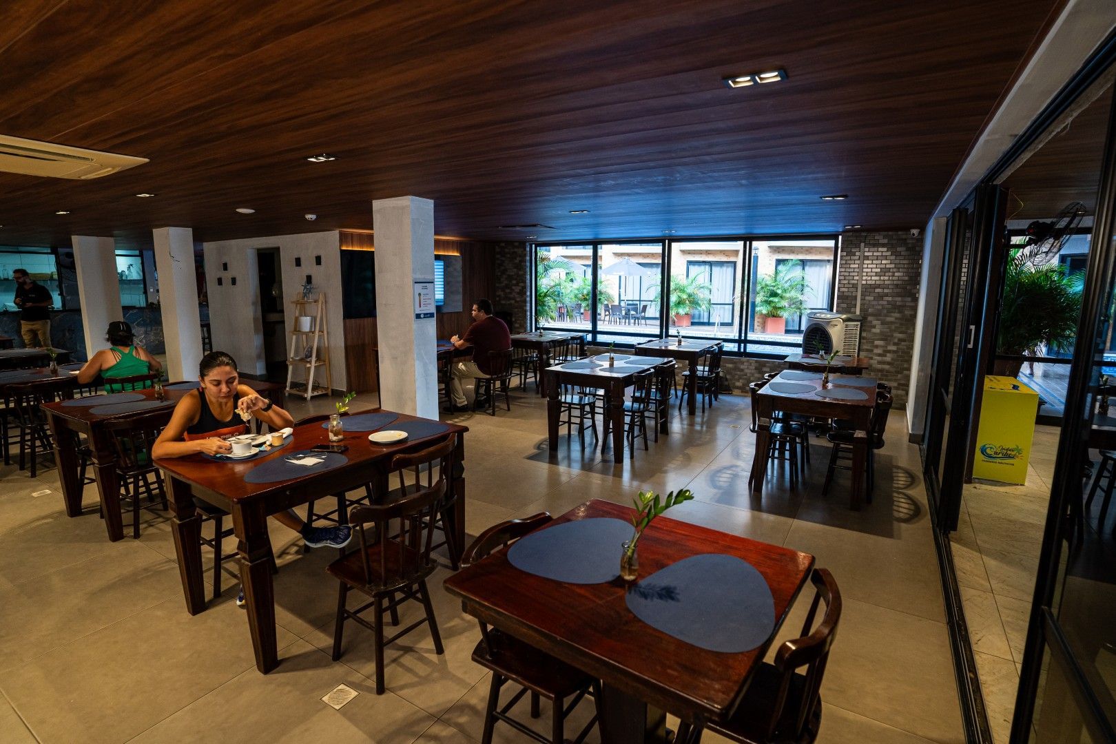 Interior espaçoso do restaurante com mesas e cadeiras de madeira, e alguns clientes perto de grandes janelas.