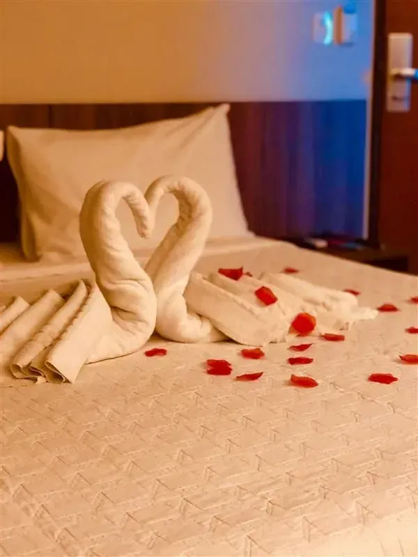 Cama de hotel romântica com cisnes de toalha em forma de coração e pétalas vermelhas espalhadas.