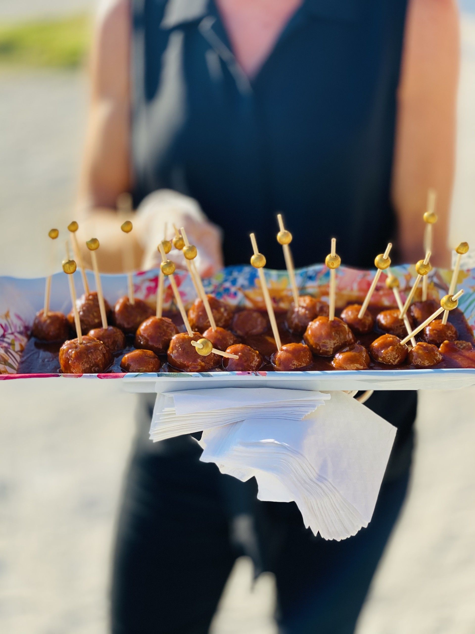 Person holding a tray of meatballs on skewers, outdoors.