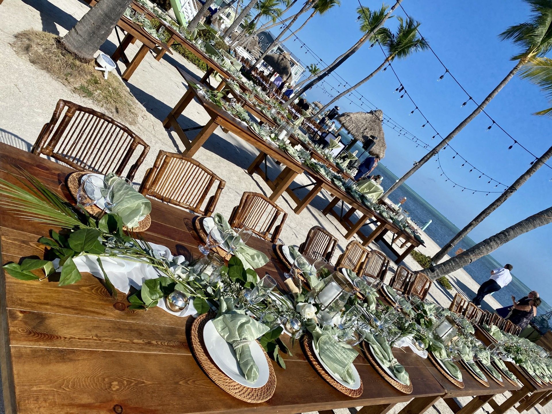 Long wooden tables set for a beachside event with floral centerpieces, woven chairs, and string lights.