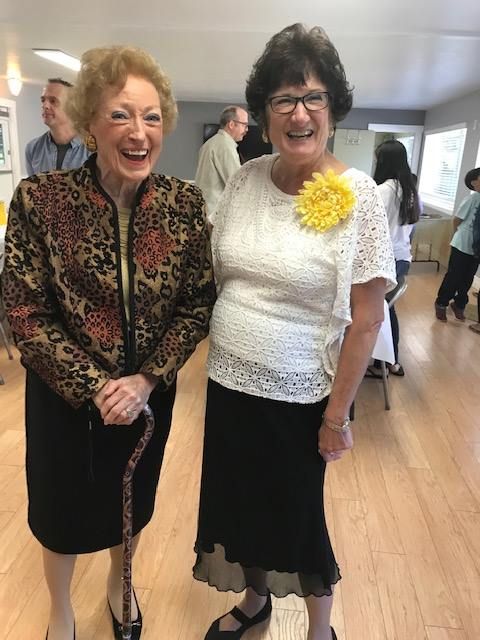 Two smiling people stand in a bright room; one wears a patterned jacket and holds a cane, the other wears a white top.