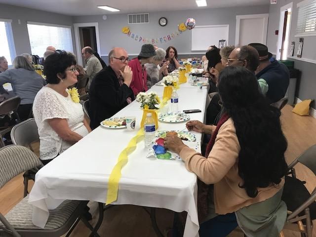 People sitting at a long table covered with a white tablecloth, decorated with yellow accents, during a social gathering.