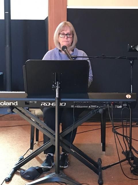 A person sits at a Roland RD-800 digital keyboard with a microphone, performing in a studio with a dark background.