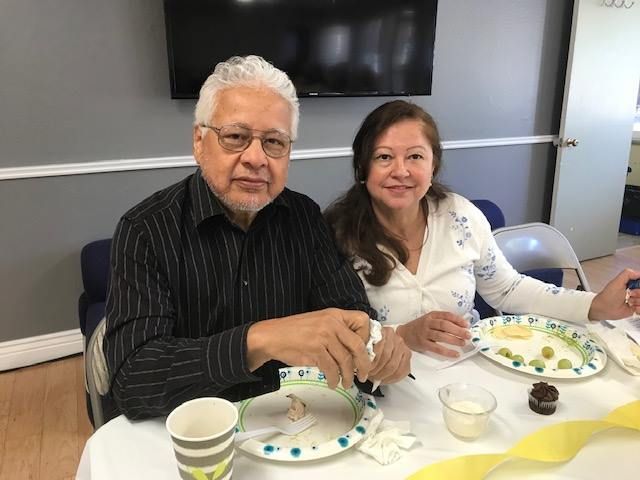 A smiling couple sits at a table with plates of food and drinks in an indoor room.