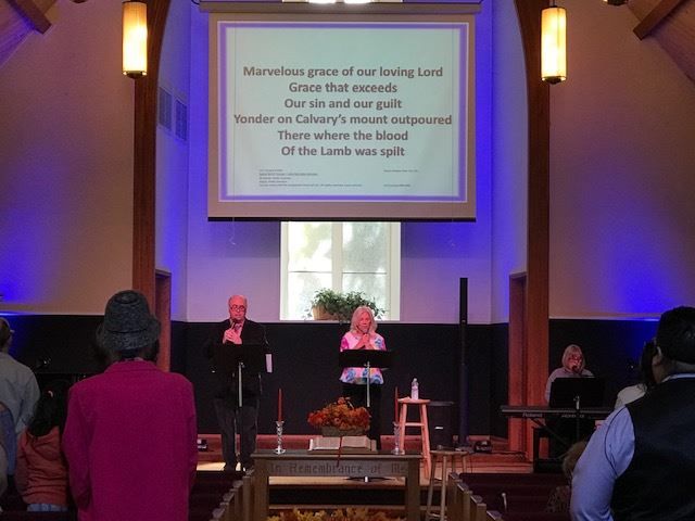 People stand at a podium in a church while lyrics are displayed on a screen above them during a service.