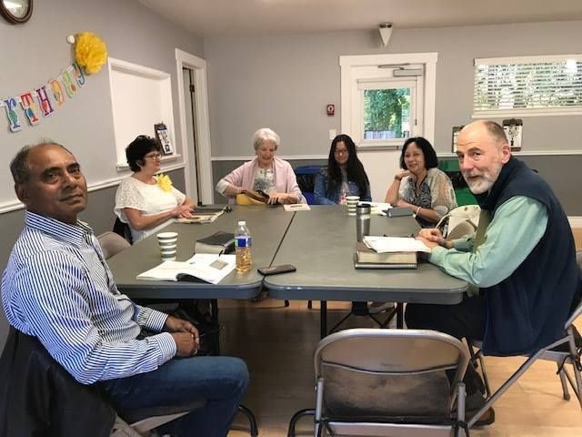 Six people sit around two joined tables in a room, appearing to participate in a meeting or study group.