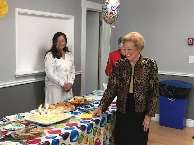 A party setting with a woman smiling near a table covered in a balloon-print cloth, a cake, and a birthday balloon.