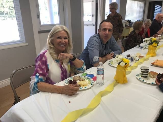 A smiling woman and a man sit at a long table set for a meal in a bright room, with yellow decorations and food.