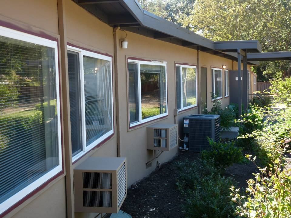 Side view of a brown one-story building with white-framed windows, window air conditioning units, and a central AC unit.