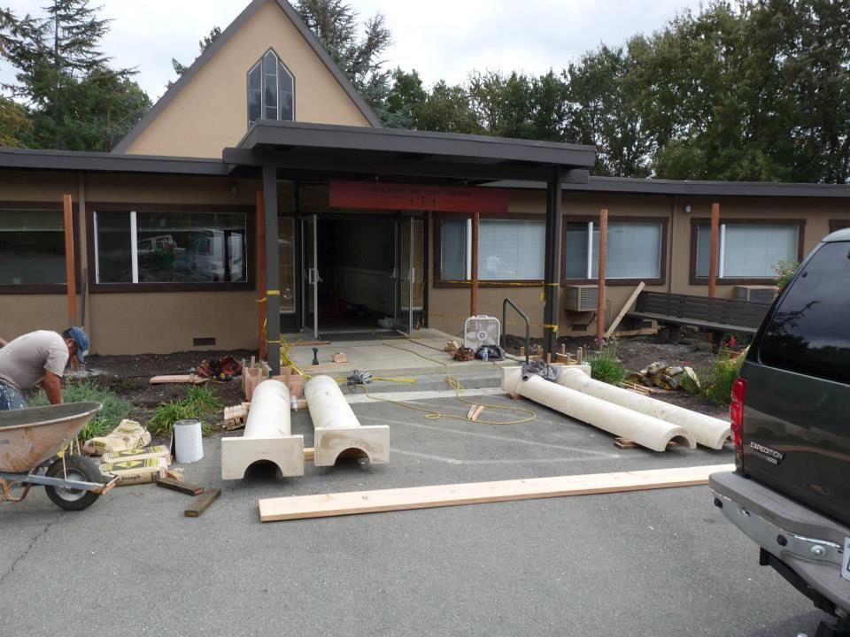 A worker and construction materials, including two white pillars, in front of a building undergoing exterior renovations.