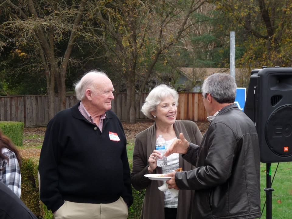 Three people talk outdoors near a large speaker, with trees and a wooden fence in the background.