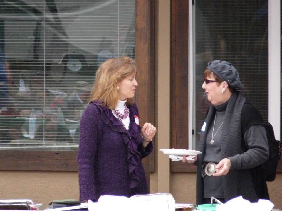 Two people stand near a building, talking while one holds a paper plate. Both wear scarves and sweaters.