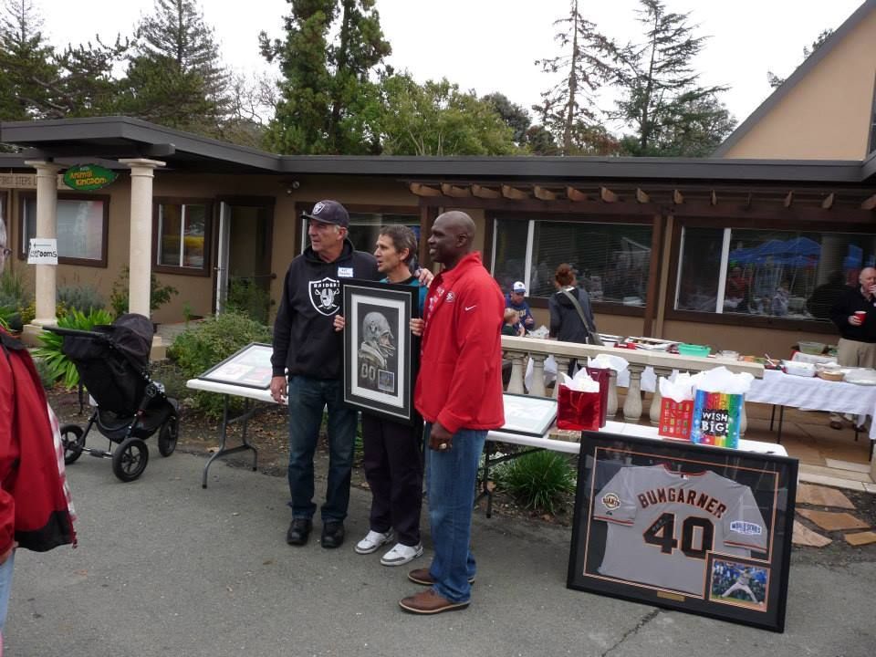 Three people stand outdoors holding a framed artwork, with a framed sports jersey on a table in front of them.