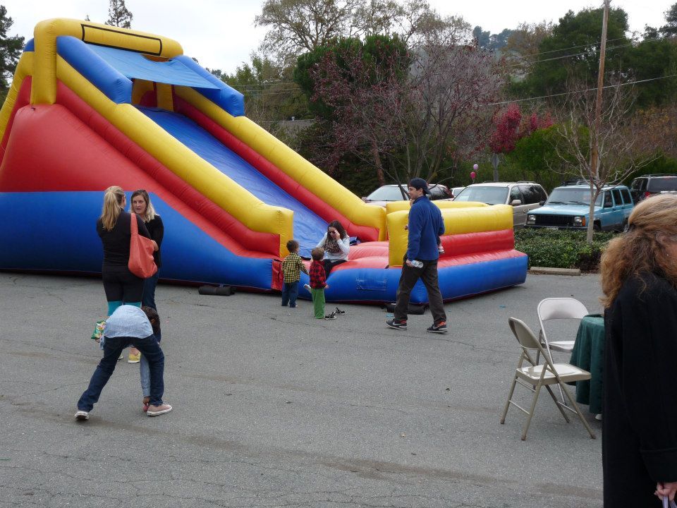 A red, blue, and yellow inflatable slide set up outdoors with people standing nearby in a parking area.