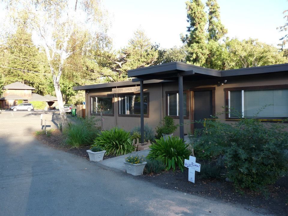 A brown single-story building with a flat roof, large windows, and a paved driveway, surrounded by trees and greenery.
