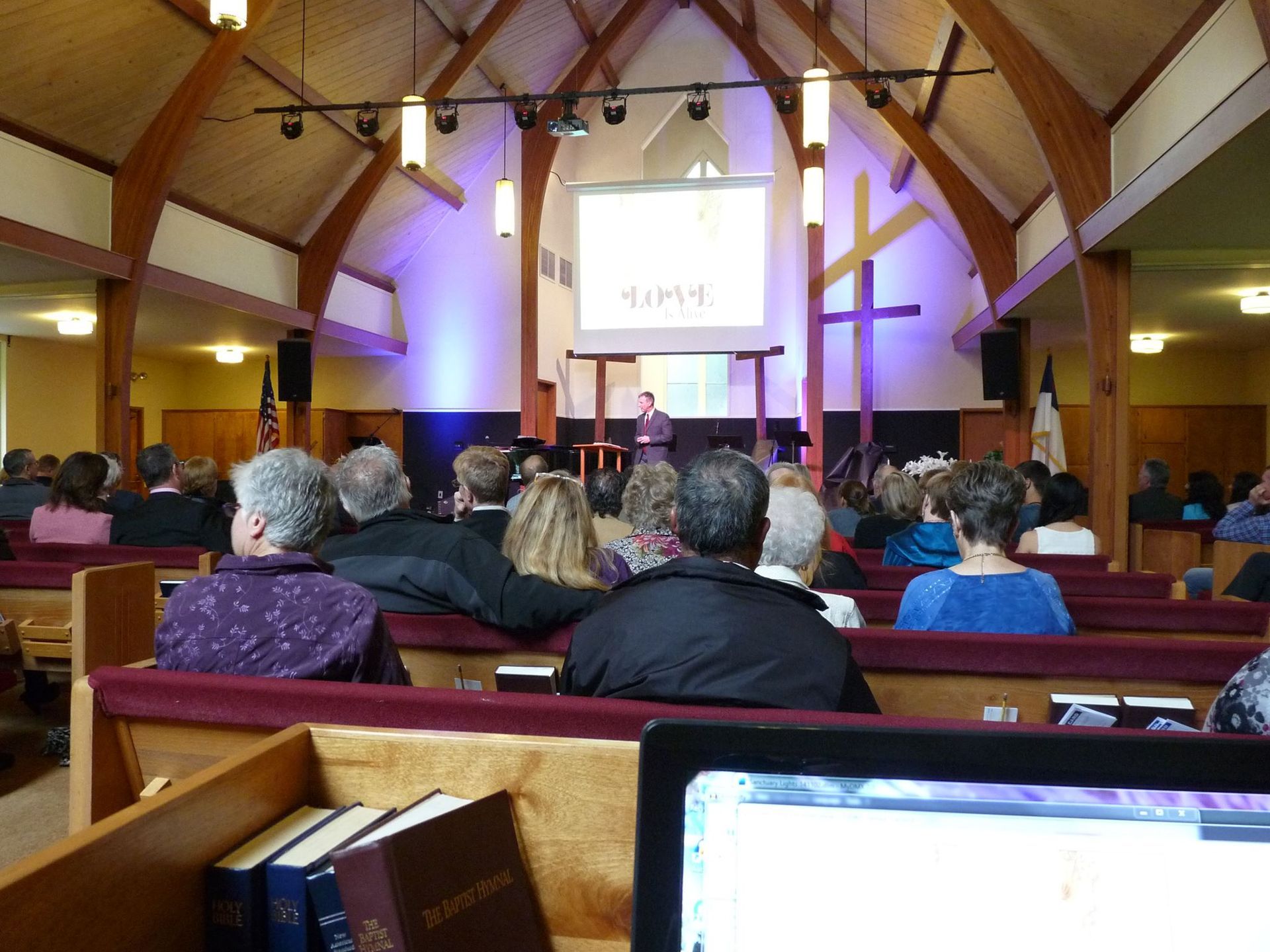 A person stands at a podium in a church sanctuary, with a congregation seated in pews facing a large screen displaying HOPE.