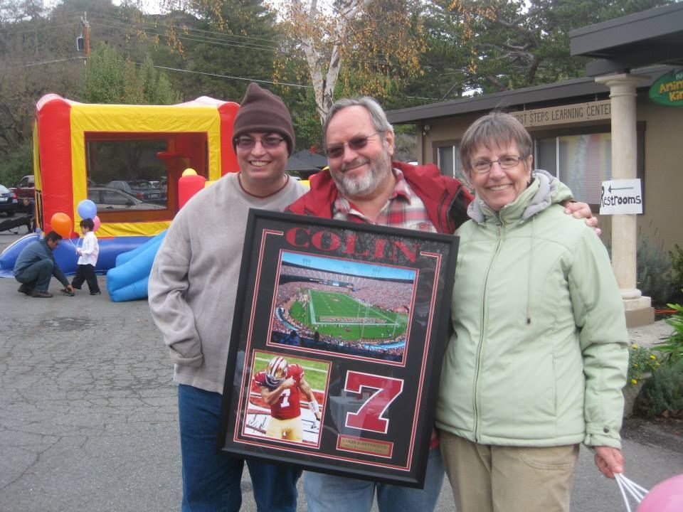 Three people smiling while holding a framed photo collage of a football stadium and player, outdoors near a bouncy castle.