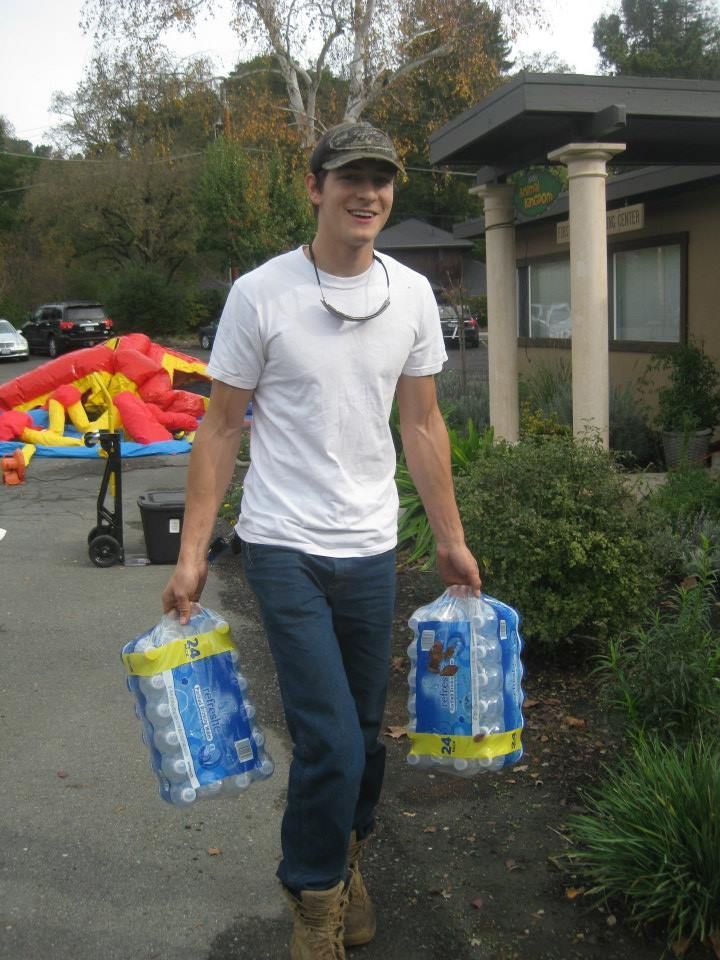 A person wearing a white t-shirt and cap walks outdoors, carrying two plastic-wrapped cases of bottled water.
