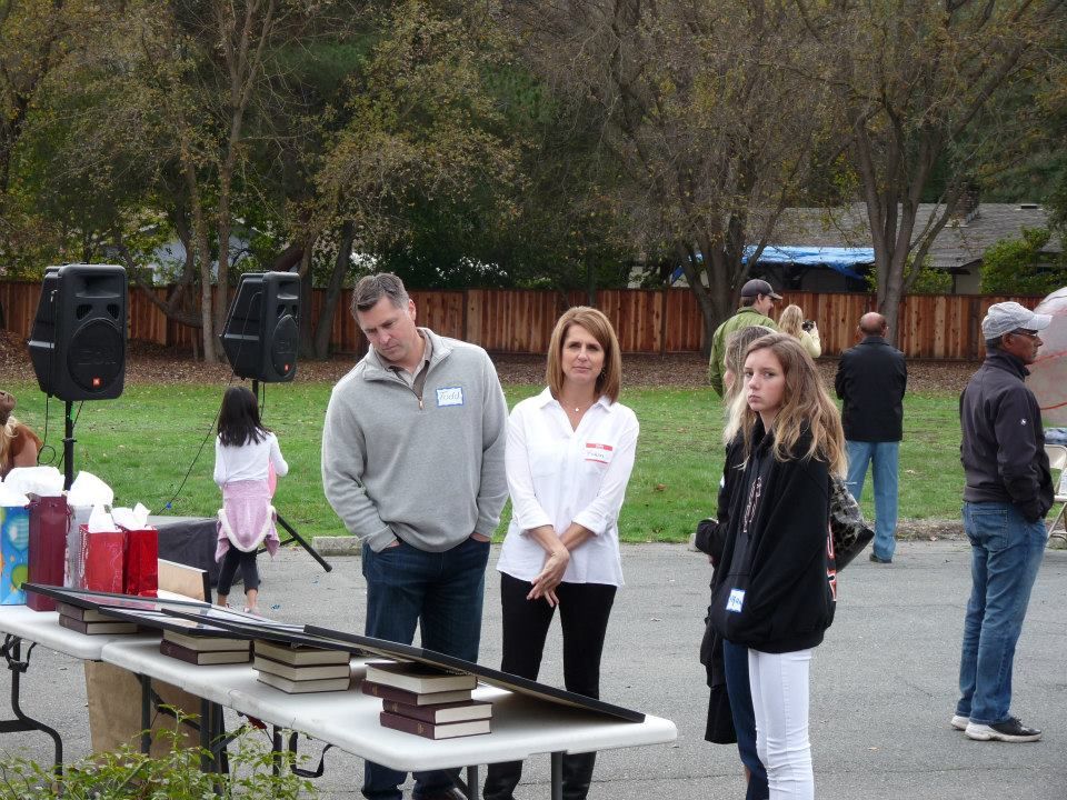 A group of people standing outside behind a table with books, with trees and a wooden fence in the background.