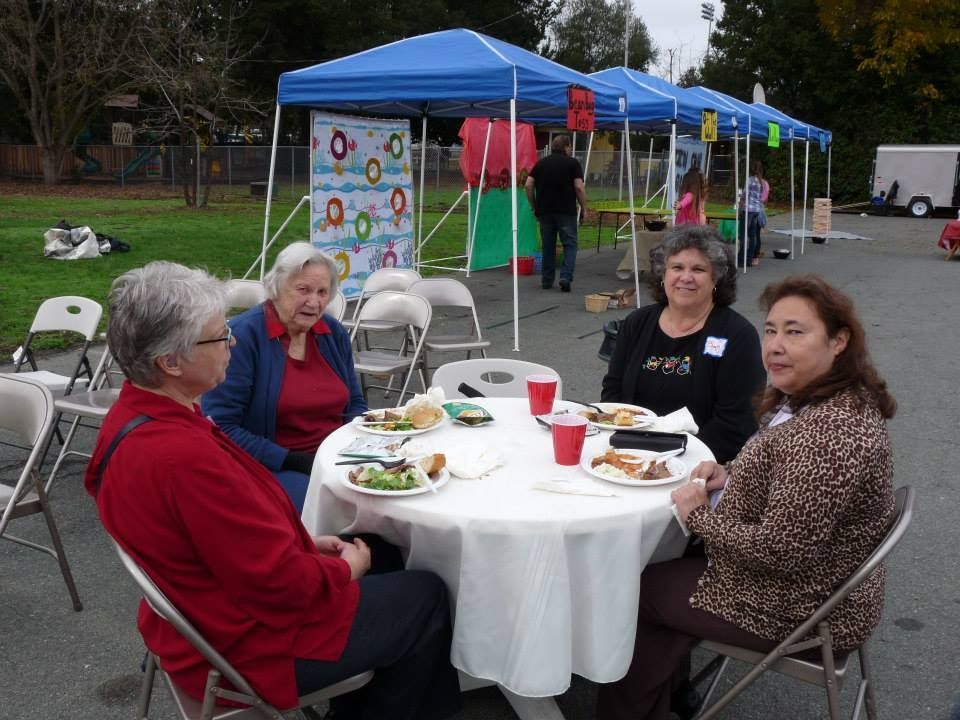 Four people eat at a round table under blue canopies at an outdoor event.