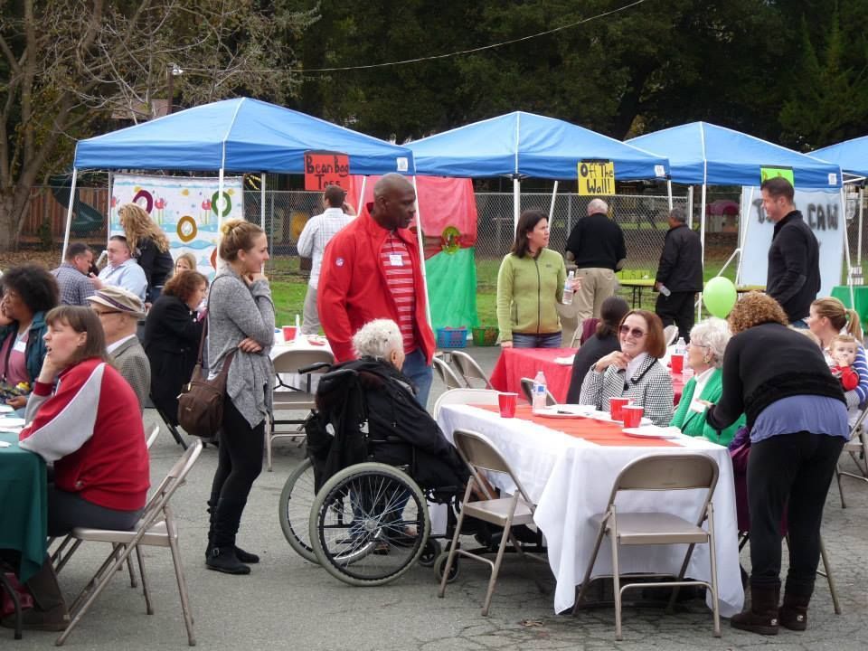 People sit and stand around tables under blue canopy tents at an outdoor social gathering or community event.