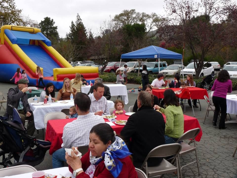 People gather at an outdoor party with a large inflatable slide and red-covered tables in a paved parking lot.