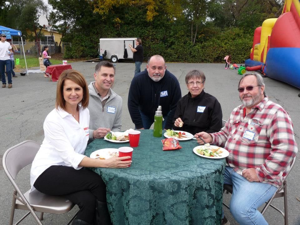 Five people sit around a table covered in a green tablecloth at an outdoor event, eating and posing for a photo.