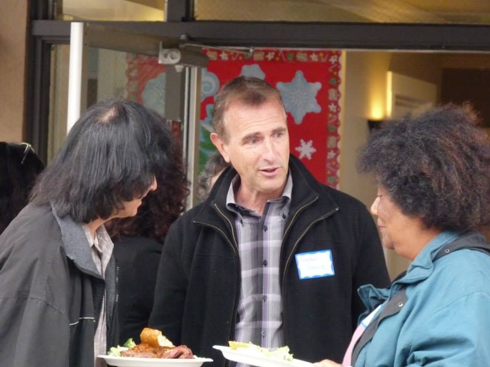 Three people stand holding plates of food at an indoor gathering, with a red decorative backdrop visible behind them.