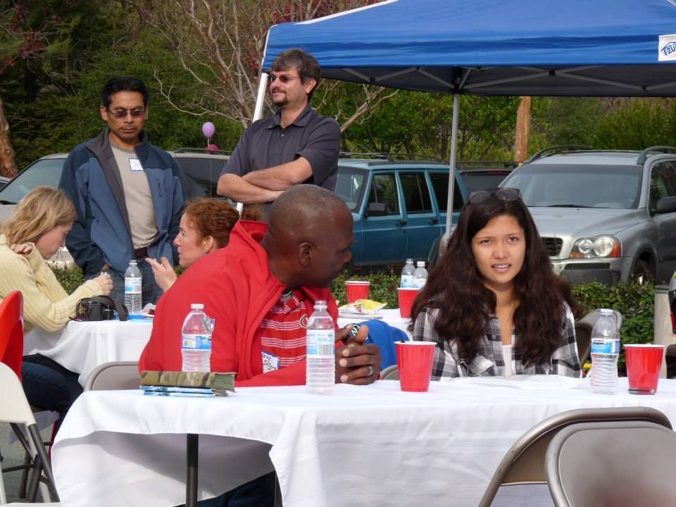 People sit at tables with drinks under an outdoor canopy at a daytime social gathering.