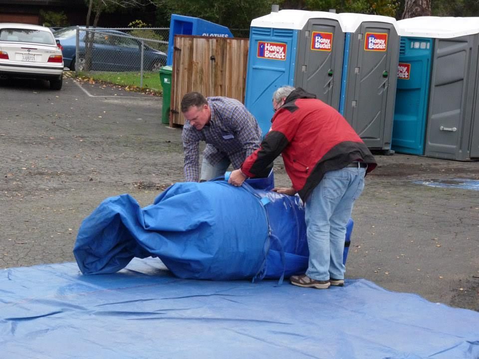 Two people in a parking lot folding a large, deflated blue inflatable structure on a blue tarp near portable toilets.